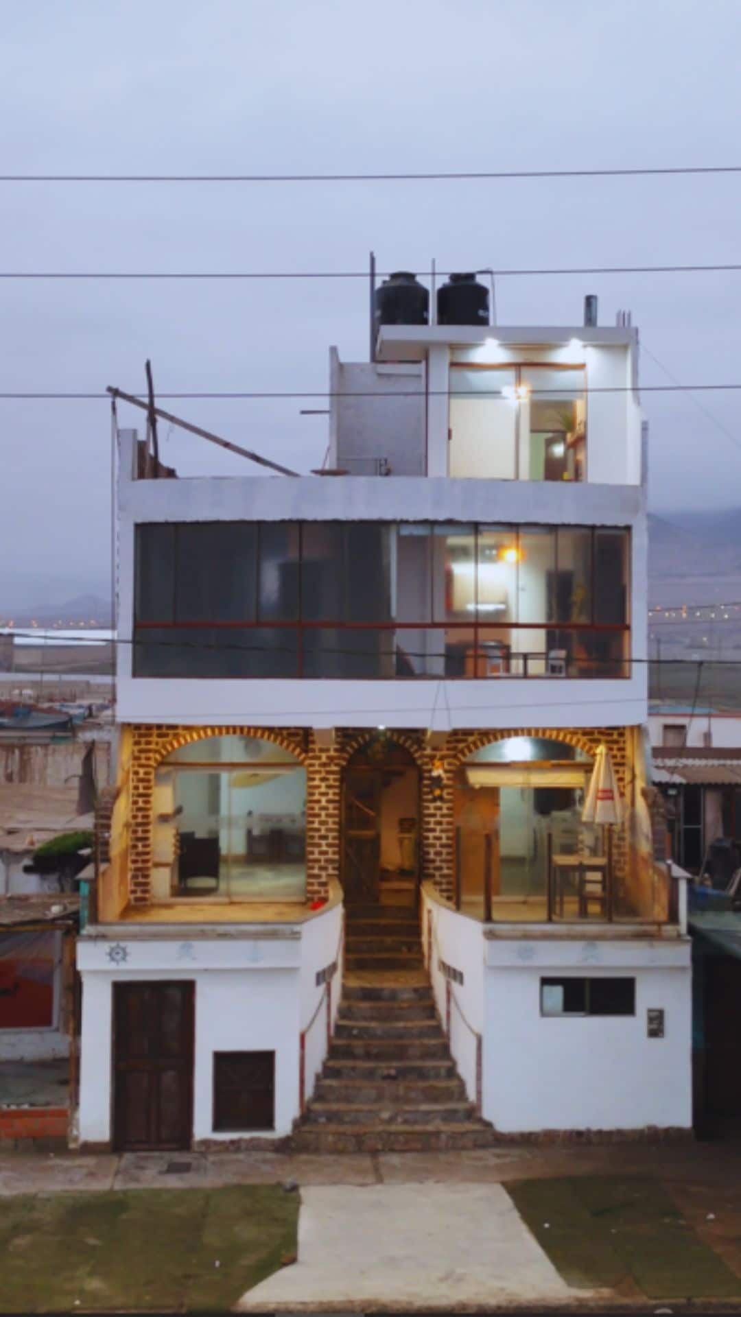 A three-story building is displayed, featuring a combination of large glass windows and a textured, patterned exterior. A staircase leads to the entrance, where warm lights are seen from within. The upper levels showcase a modern design with outdoor spaces, reflecting a coastal atmosphere.