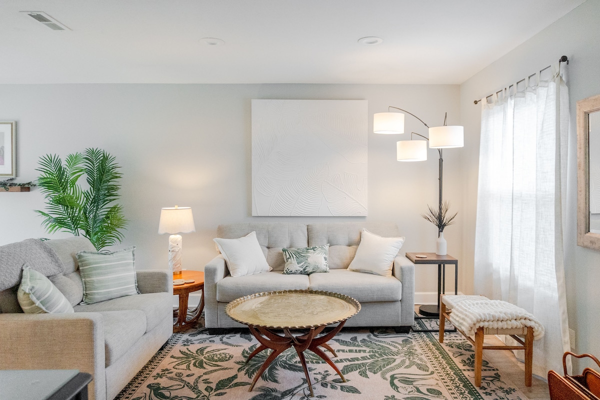 A cozy living area features a light-colored sofa and a loveseat, paired with a round, wooden coffee table. A floor lamp and a table lamp provide soft lighting. Sheer curtains frame a window, while a decorative plant and art piece add subtle accents to the space.