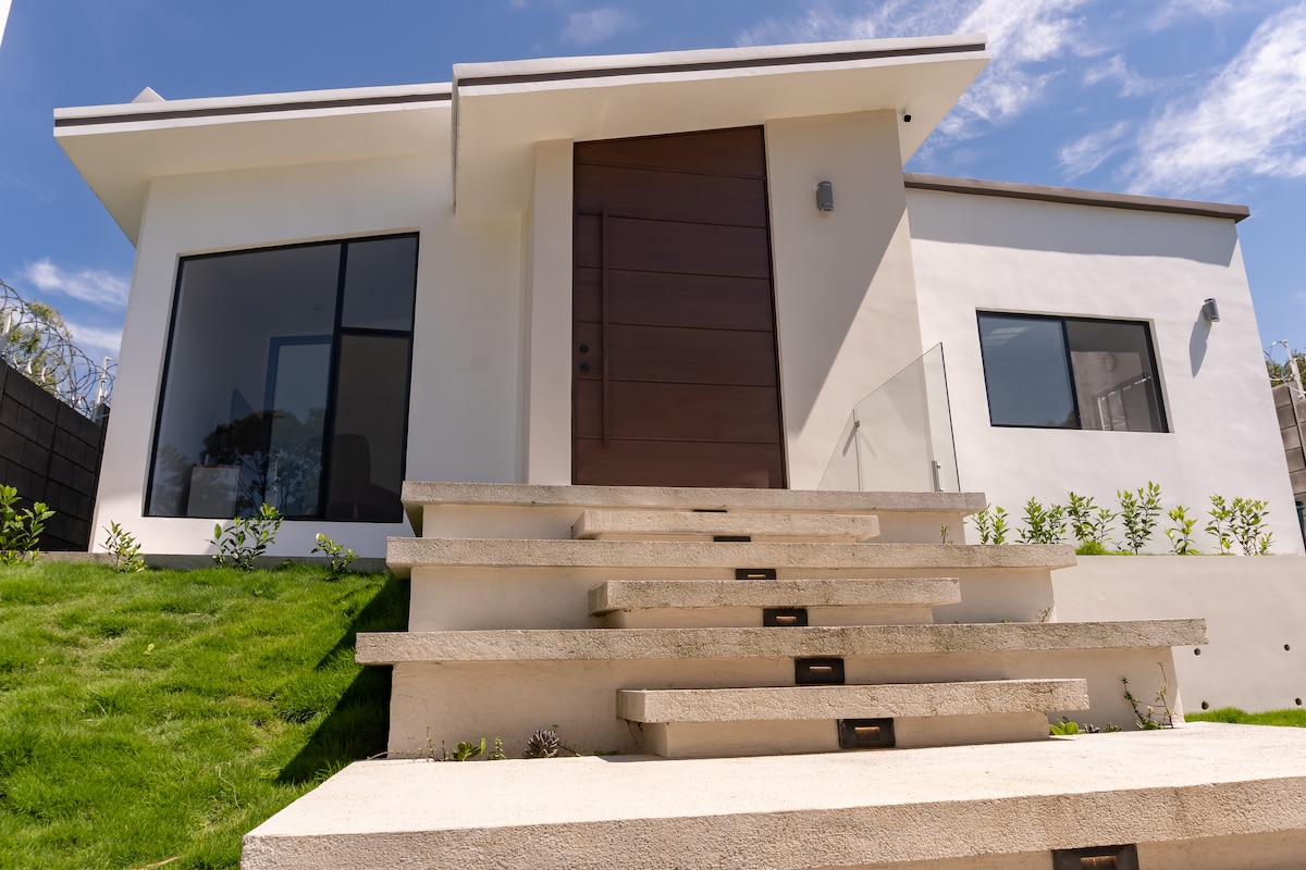 The exterior of a modern home is presented, featuring a sleek wooden front door. Stone steps lead up to the entrance, bordered by neatly trimmed grass and small shrubs. Large windows on either side of the door enhance natural light, contributing to the home’s contemporary aesthetic.