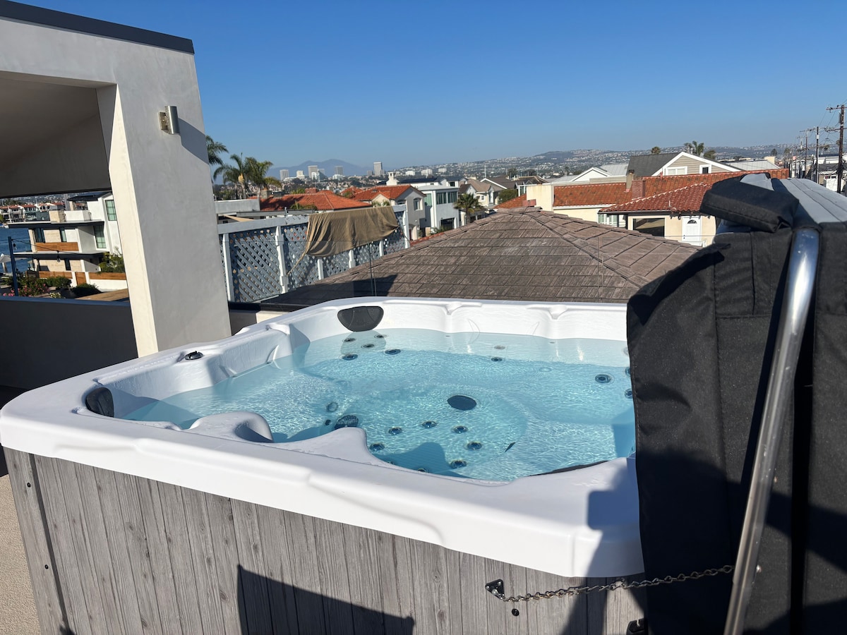 A spacious Jacuzzi tub is situated on the rooftop patio, offering panoramic views of the surrounding area. The hot tub features multiple jets and is surrounded by a wooden enclosure. Clear skies and distant city views enhance the serene environment.