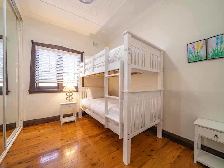 Chambre ️ confortable avec lits superposés en bois blanc, draps frais et plancher en bois poli✨. Comprend deux tables de chevet avec des lampes uniques, une armoire miroir et de l'art mural pour une touche chaleureuse.