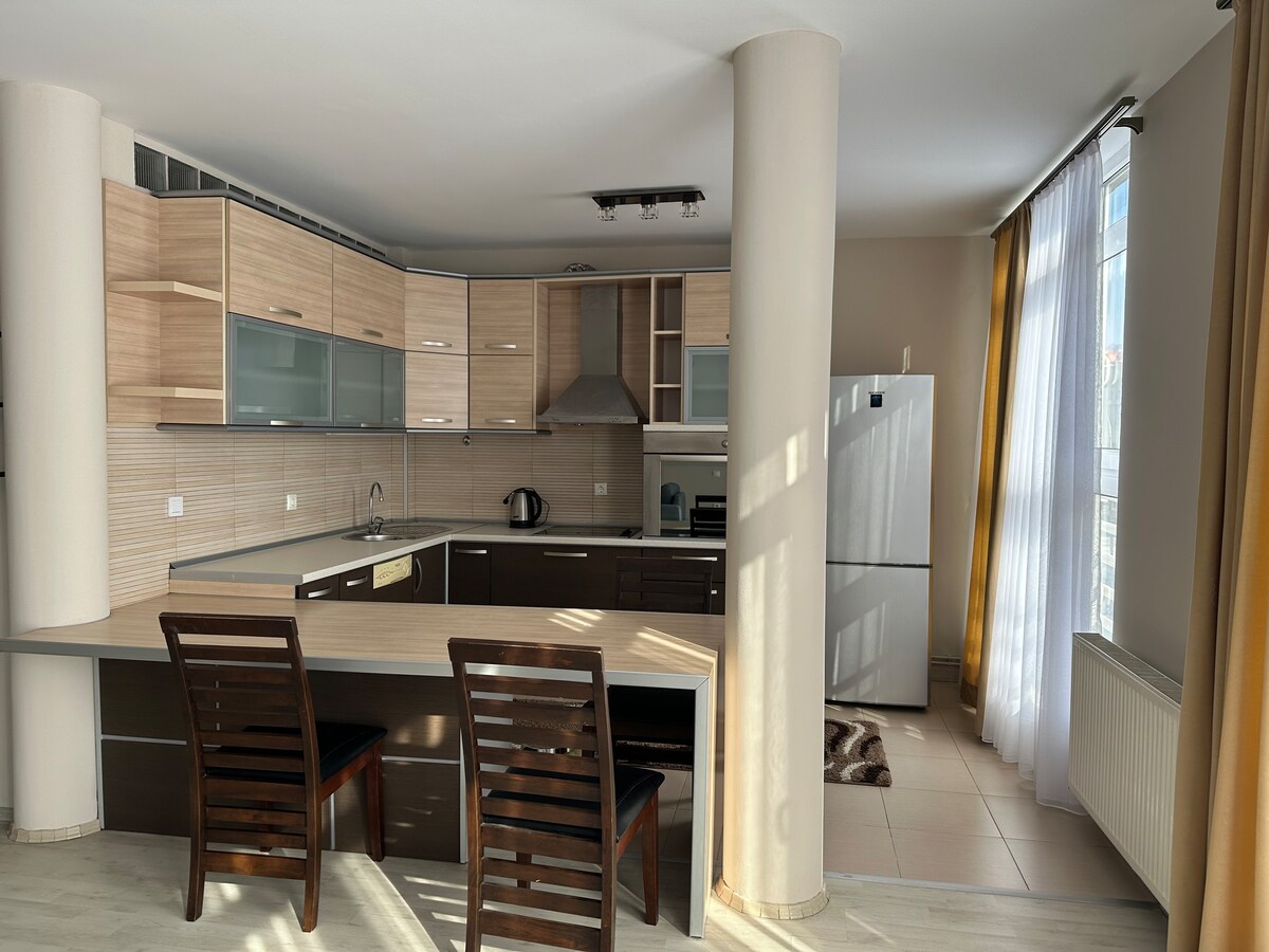 A modern kitchen space features sleek cabinetry and an island with two wooden chairs. Natural light pours in through large windows, illuminating the countertops and a white refrigerator. Essential appliances are visible, contributing to a fully equipped cooking area.