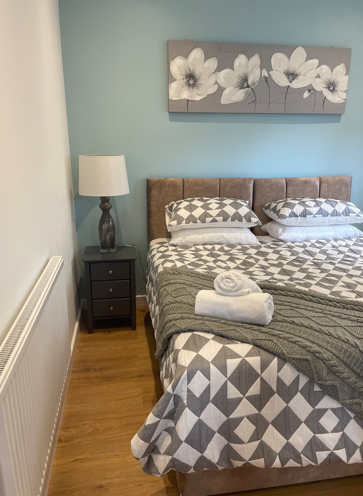 A modern bedroom features a spacious bed with geometric patterned bedding and plush pillows. A bedside table with a lamp provides soft lighting. A radiator is visible along one wall, and a floral wall art piece adds a touch of decor to the serene blue background.