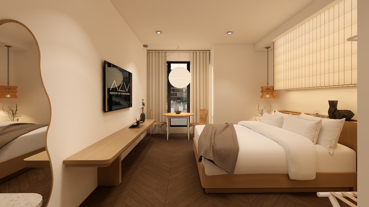A minimalist studio features a spacious King-sized bed, complemented by soft textures and neutral tones. A wooden dining table is positioned near a window, providing natural light. A television is mounted on the wall opposite the bed, and a decorative piece rests on the bedside table.
