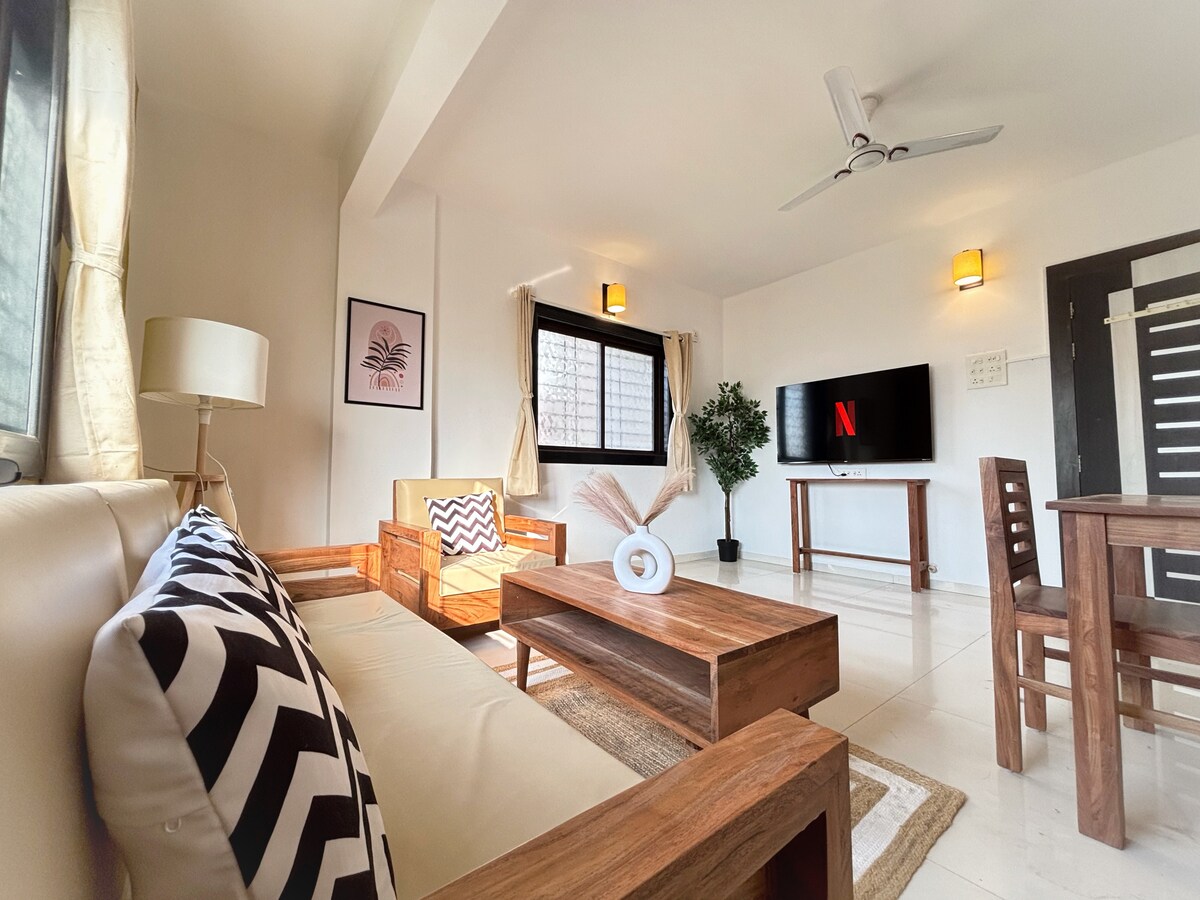 A well-lit living area features a comfortable cream-colored sofa adorned with patterned cushions. A wooden coffee table sits centrally, accompanied by a small dining table in the background. A flat-screen TV is mounted on the wall, with decorative plants adding a touch of greenery.