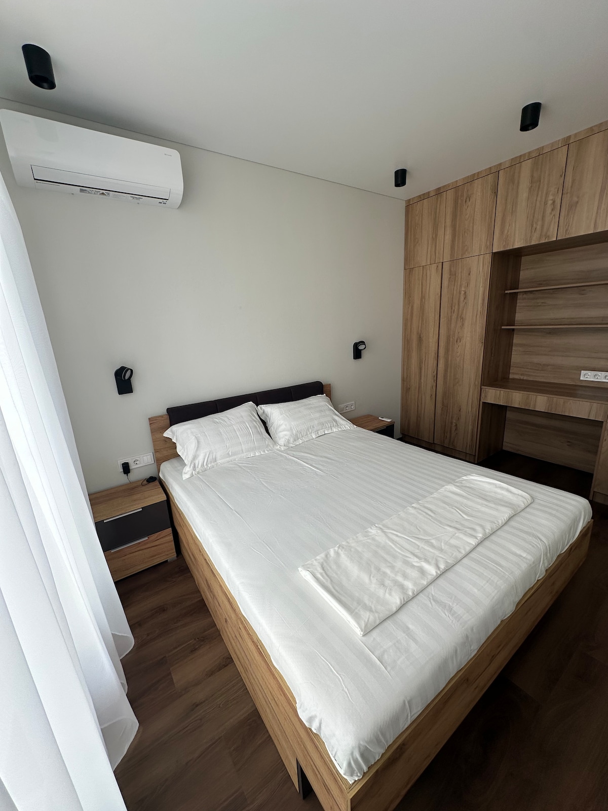 The bedroom features a spacious bed with a light-colored duvet and decorative pillows, set against a neutral wall. Warm wooden accents are seen in the furniture, including a bedside table and built-in shelves. Natural light enters through a sheer curtain-covered window, enhancing the airy feel.