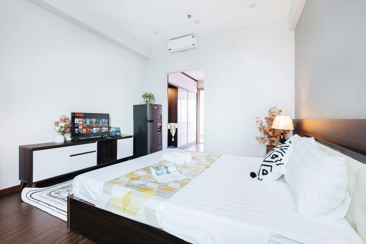 A modern sleeping area features a queen-sized bed dressed in white linens and a patterned throw. A flat-screen television is positioned on a low cabinet, while a mini refrigerator stands nearby. Natural light filters through a spacious doorway to another area of the home.