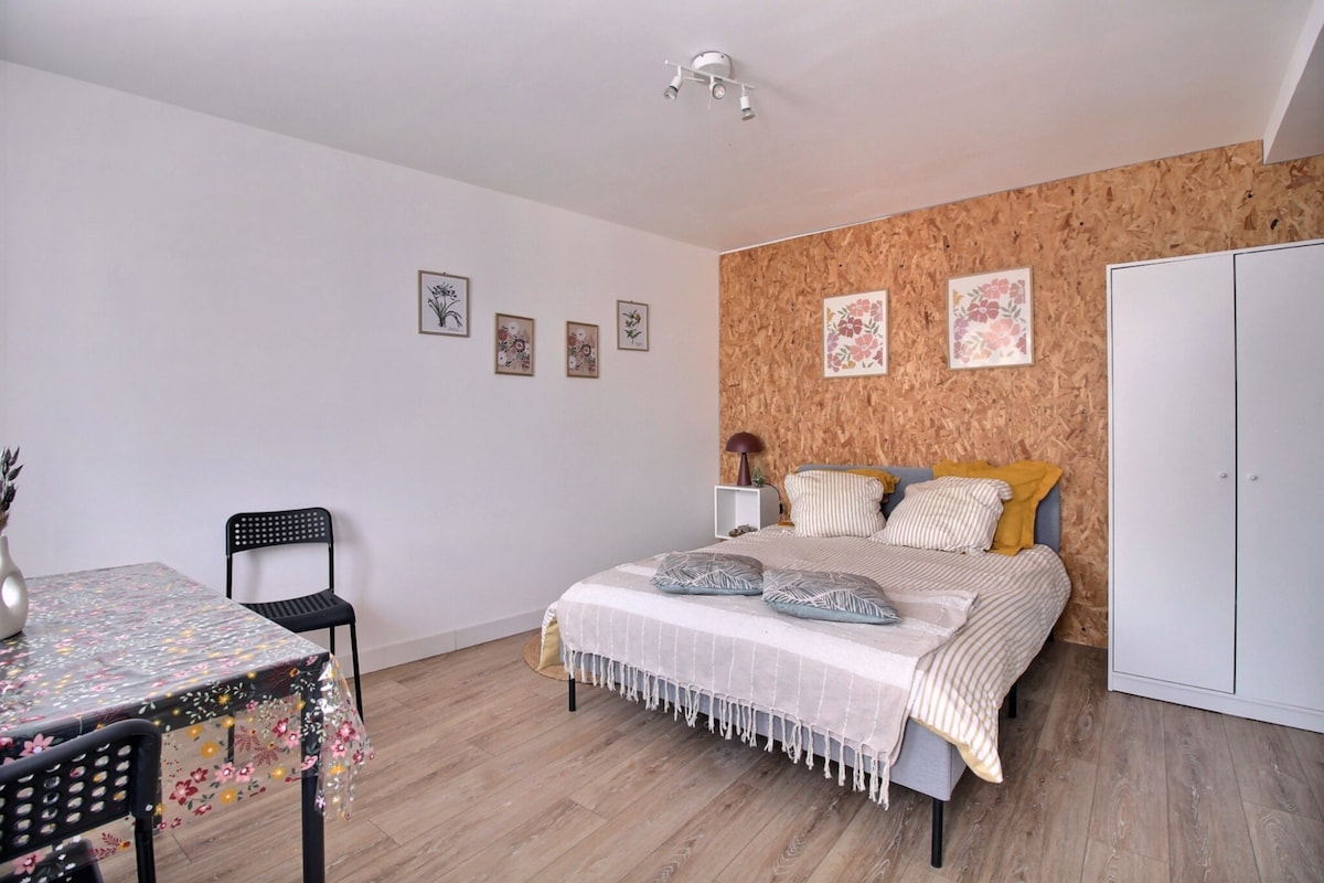 The spacious studio features a comfortable bed dressed in fresh linens and decorative pillows. A simple table with a floral tablecloth is positioned nearby, accompanied by two black chairs. Light-colored walls and a textured wood accent wall create a soothing ambiance.