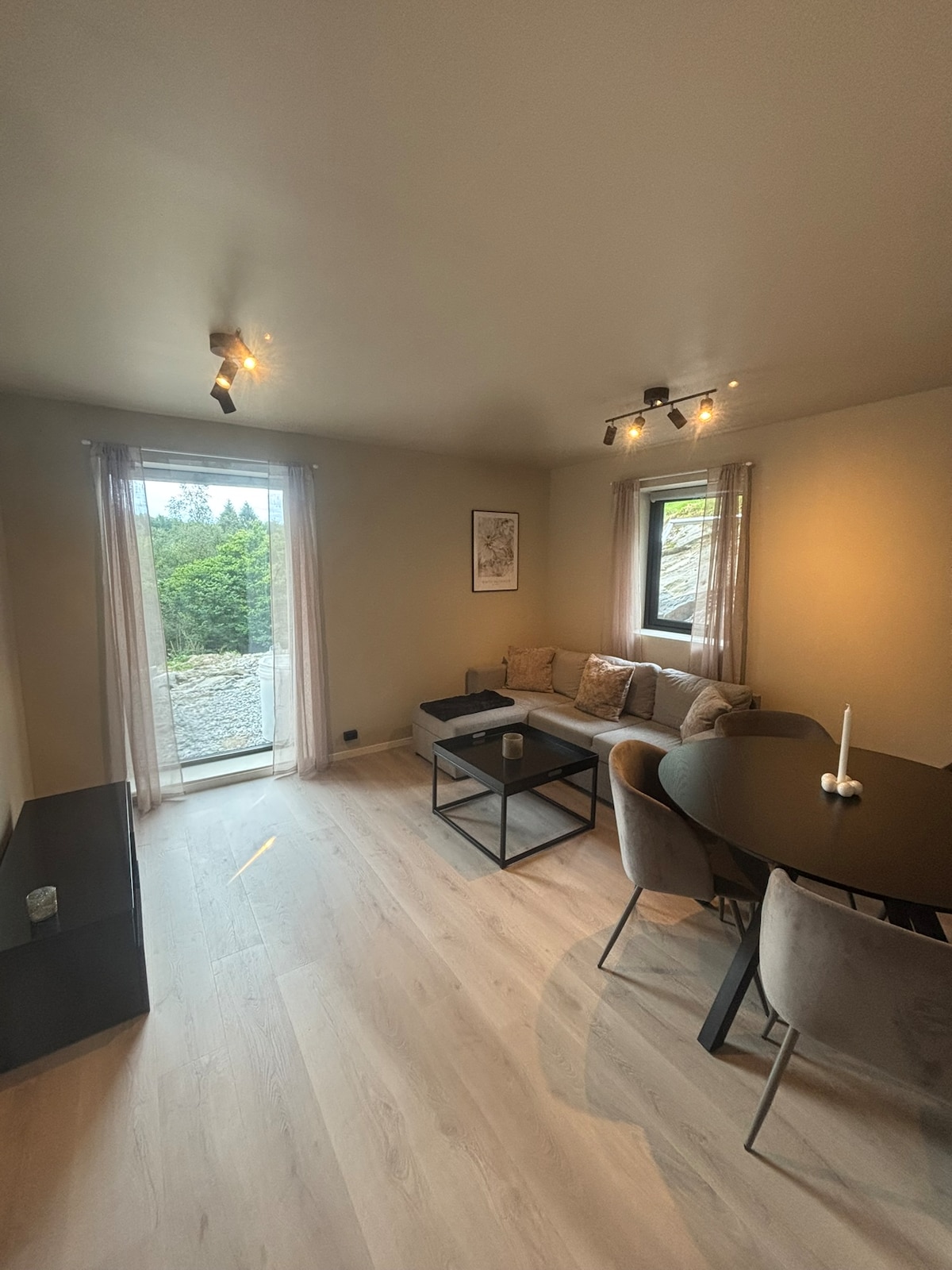 A well-lit living area is presented, featuring a modern seating arrangement with a cozy sofa and a coffee table. A round dining table with several chairs is positioned nearby. Large windows allow natural light to fill the space, offering views of the surrounding greenery.