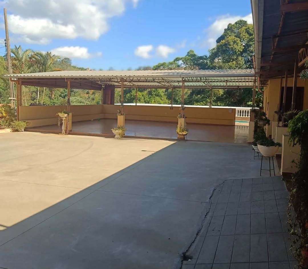 A spacious outdoor area is presented, featuring a large concrete surface surrounded by greenery. The covered section offers shade, with potted plants positioned along the edges, creating an inviting space suitable for gatherings and relaxation.