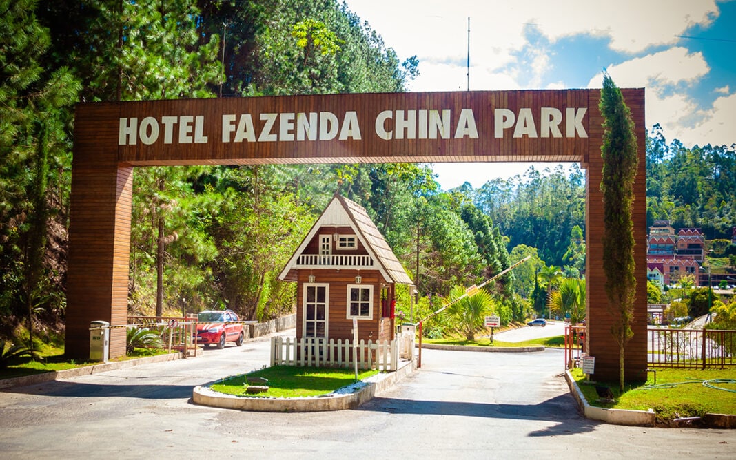 An entrance archway welcomes guests to Hotel Fazenda China Park. The wooden structure features the hotel name prominently displayed, flanked by tall trees and a small replica house. A clear sky and lush greenery frame the scene, indicating a serene environment.