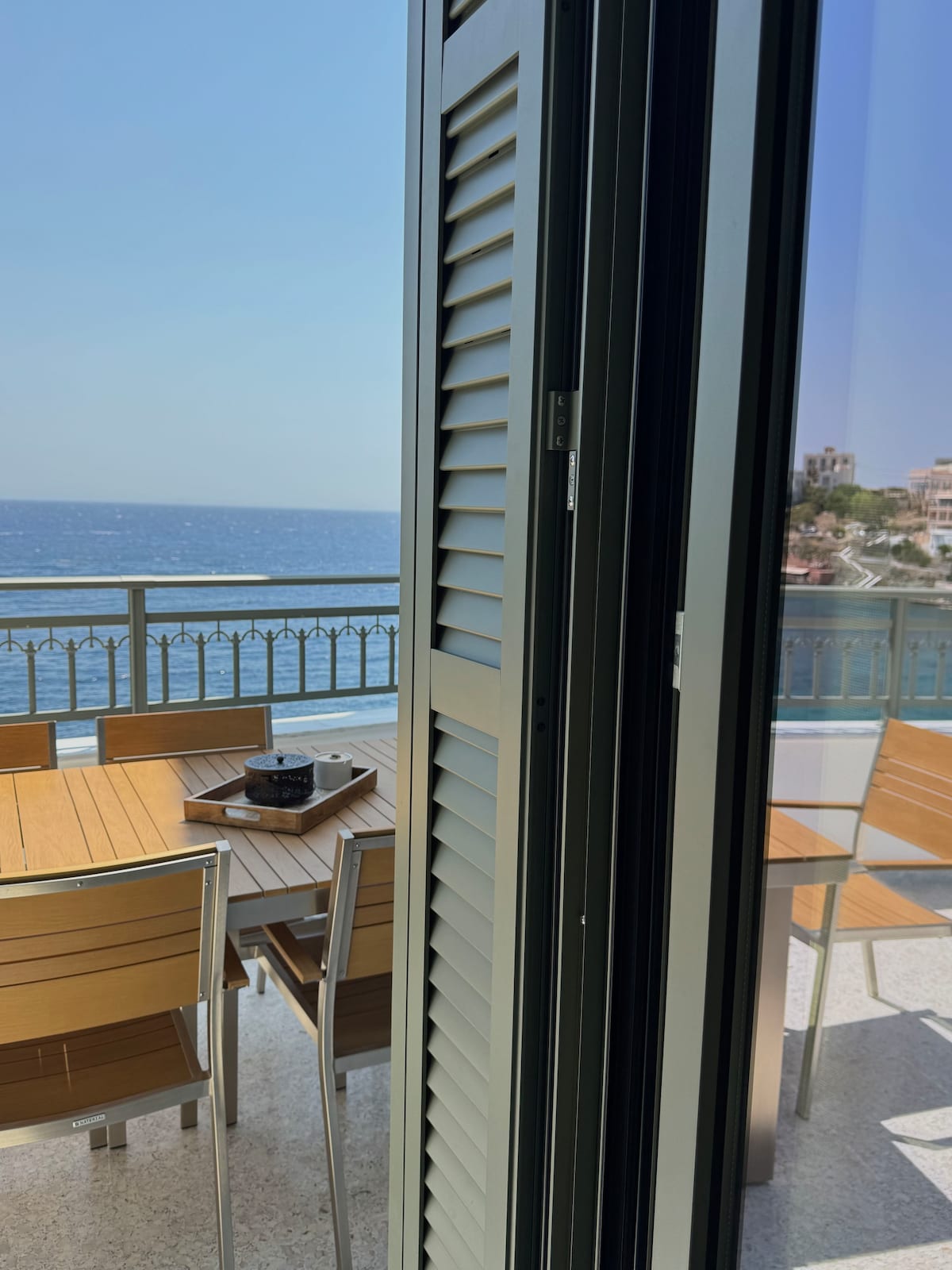 A view of a spacious terrace is captured through open shutters, showcasing a wooden dining table surrounded by metal chairs. The blue sea glistens in the background, complemented by a clear sky, enhancing the connection between indoor and outdoor spaces.