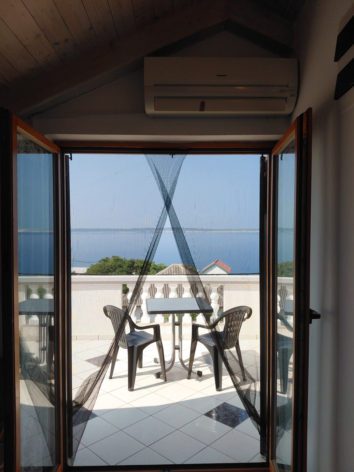 A view of a balcony area is framed by open double doors, showcasing a small table set with two chairs. An expanse of water is visible in the distance, with a clear sky complementing the tranquil scenery. Light tile flooring enhances the airy atmosphere.