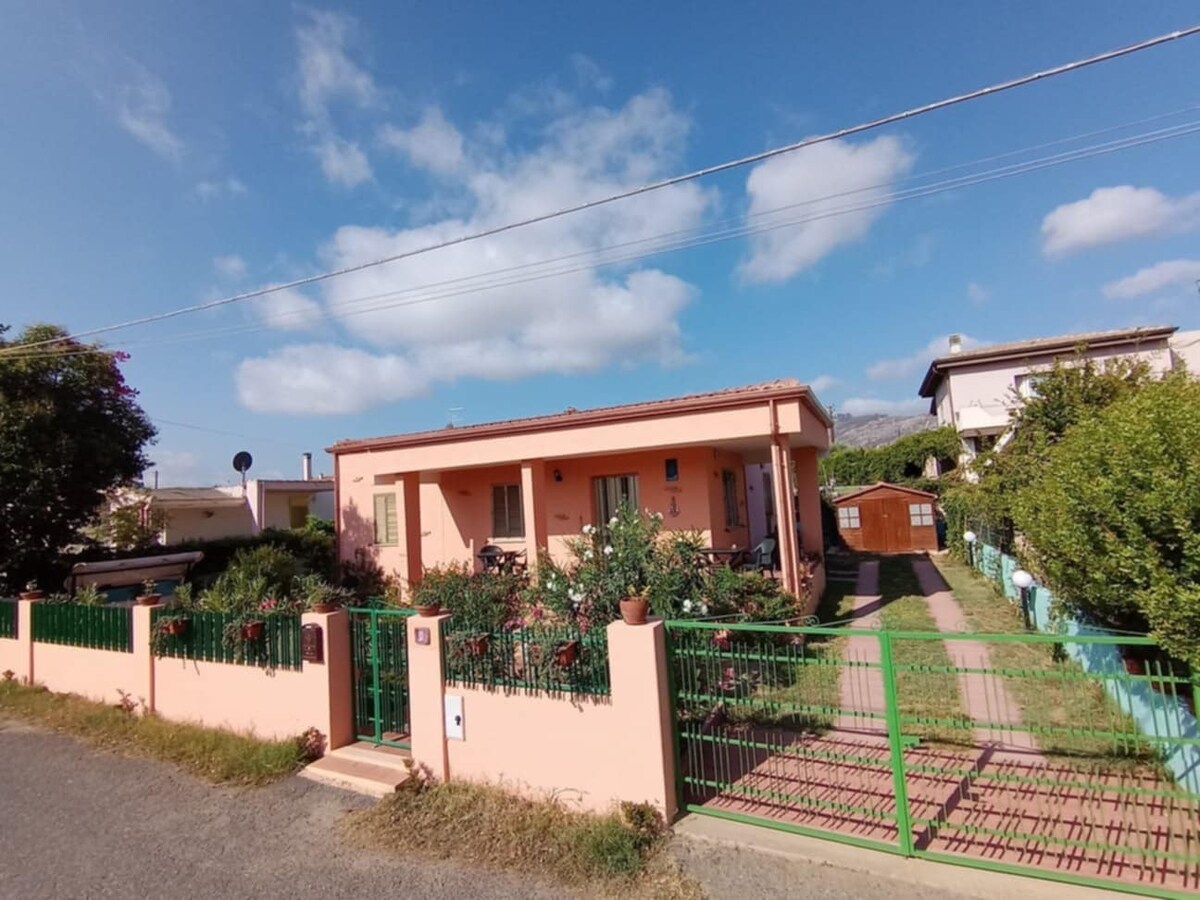 A charming villa is set behind a green fence, surrounded by vibrant vegetation. The single-story structure features a warm pink exterior and a welcoming entrance. A pathway leads through the garden, offering a glimpse of the nearby residential area and blue sky above.