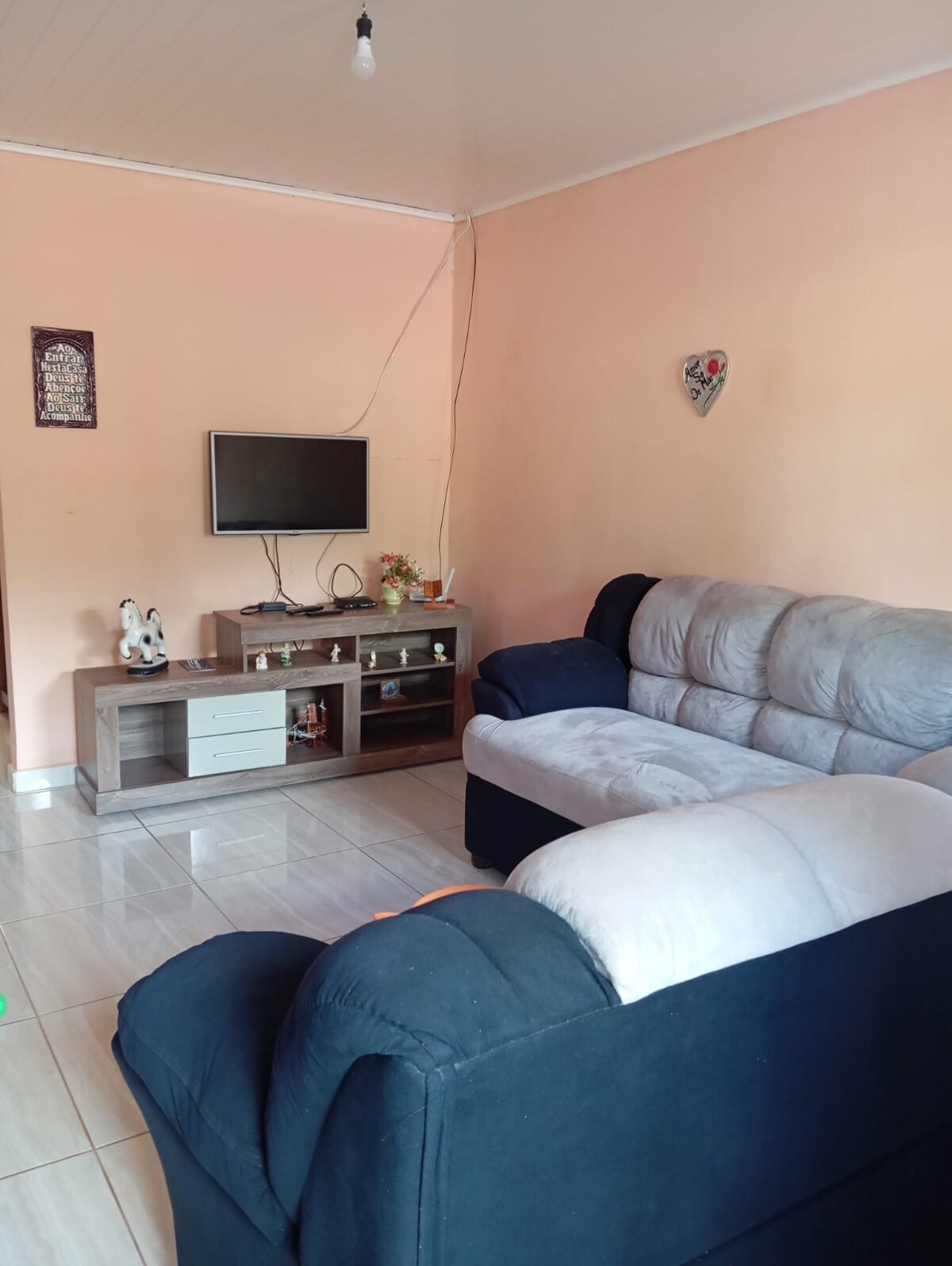 A comfortable living area is presented, featuring a dark blue armchair and a light gray sofa. A simple entertainment unit is placed against a peach-colored wall, supporting a flat-screen television and decorative items. The floor is tiled with a glossy finish, enhancing the room's spacious feel.
