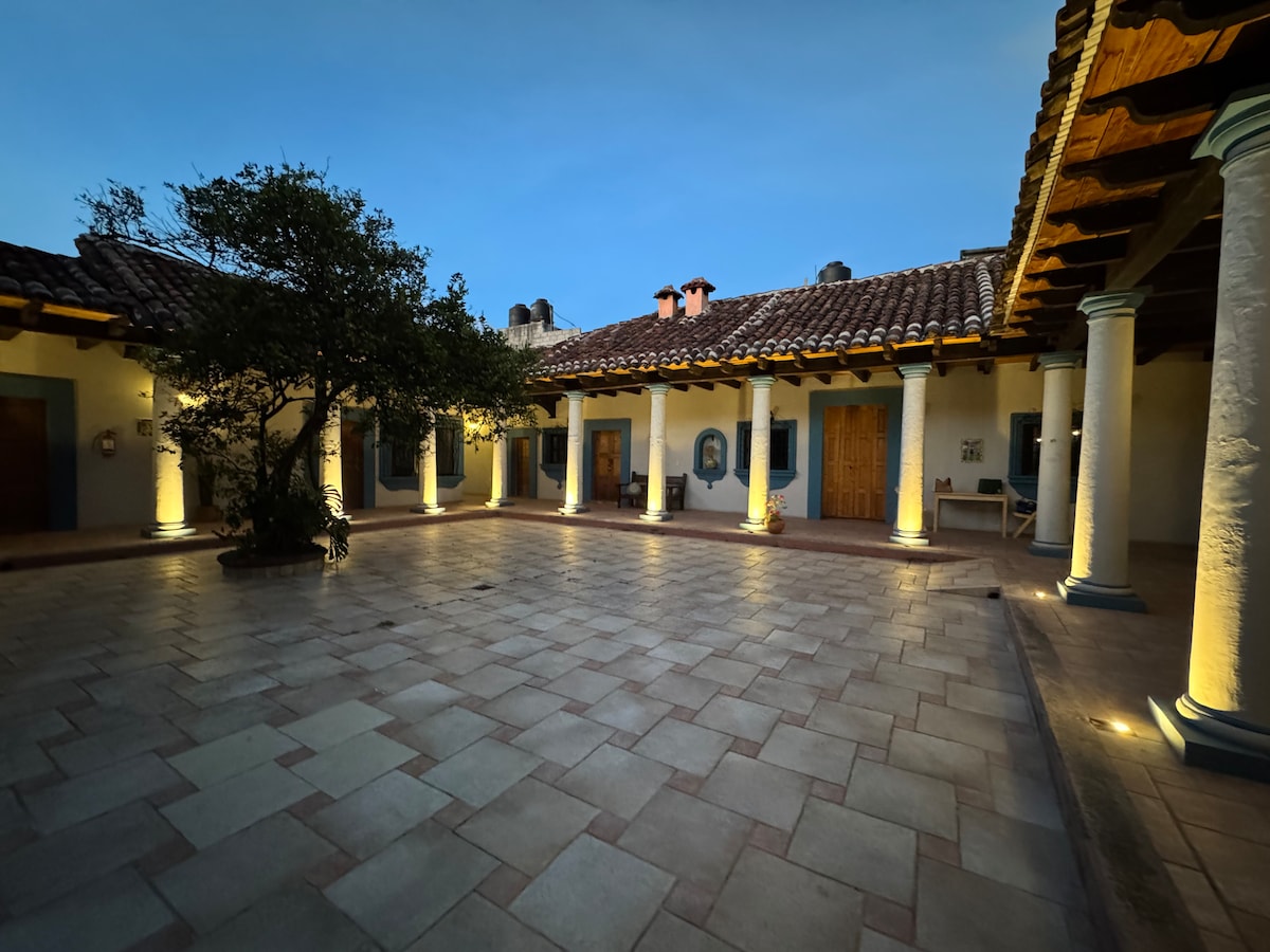 A serene courtyard is illuminated by soft lighting, highlighting textured stone tiles and wooden beams. Tall columns surround the space, while lush greenery is featured in the center, creating a tranquil environment that invites relaxation.