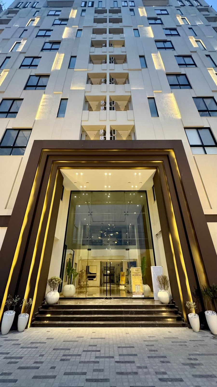 The building entrance is marked by a modern façade featuring a series of illuminated geometric patterns. Large glass doors provide access to the lobby area, while elegant potted plants flank the entrance steps, creating a welcoming approach.