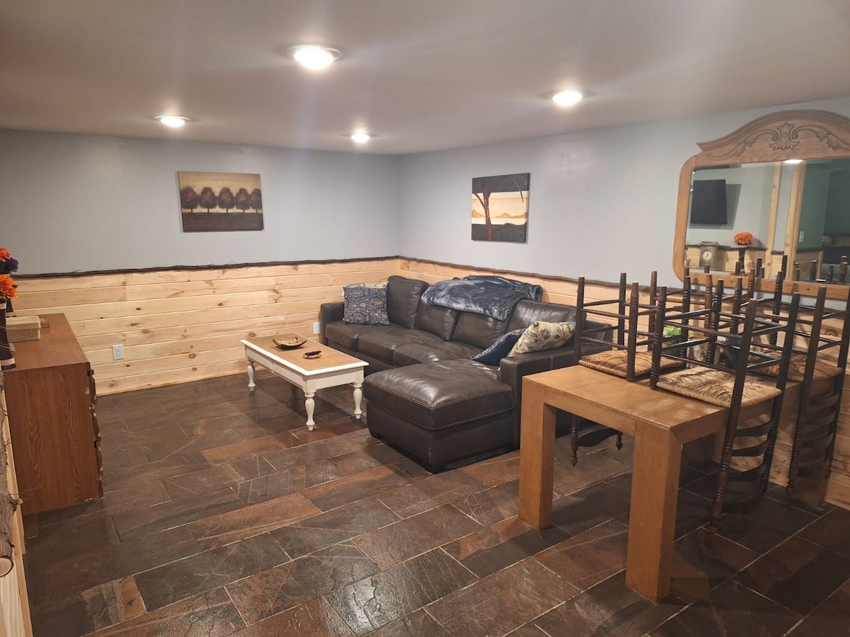 The cozy living area is framed by wood-paneled walls, featuring a dark leather sofa and a light coffee table. A wooden dining table with stacked chairs sits nearby, while art pieces adorn the walls. Warm lighting enhances the earthy stone flooring.