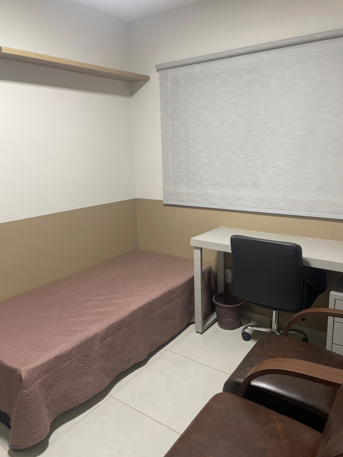 A modestly furnished room features a single bed draped with a light brown cover. A small desk with a black chair is positioned beside the bed. Natural light filters through a window covered by a light shade, and a potted plant adds a touch of greenery.