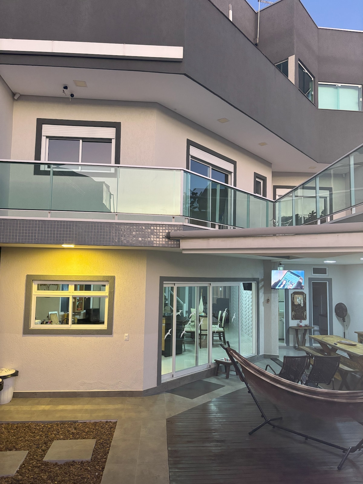 The image displays a modern triplex structure with multiple levels and large glass windows. A spacious outdoor area is visible, featuring a wooden deck and a relaxing hammock. Indoor dining space is showcased through the glass doors, blending indoor and outdoor living seamlessly.