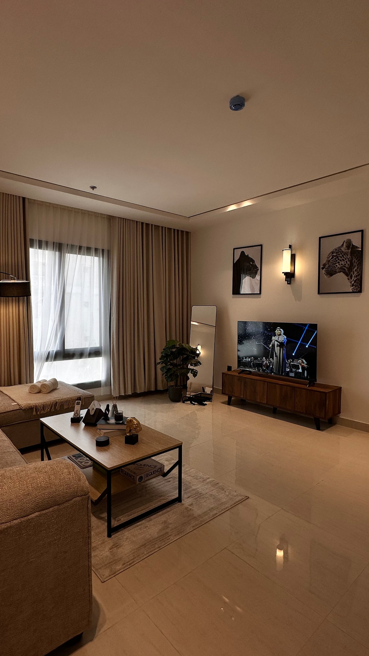 A comfortable living area features a light-colored sofa paired with a stylish wooden coffee table. Neutral-toned curtains drape beside large windows, allowing natural light to filter in. Stylish wall art depicting horses complements the modern decor, while a floor mirror adds depth to the space.