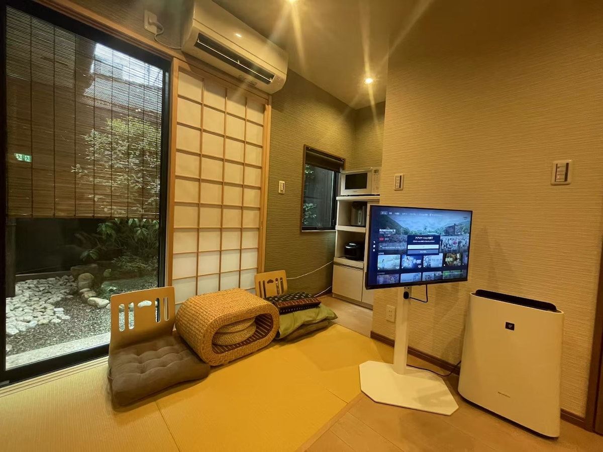 A cozy living area features a tatami mat with a light yellow color and comfortable cushions arranged on it. A small traditional garden is visible through the large glass windows, along with a television on a stand. A compact kitchenette is also present.