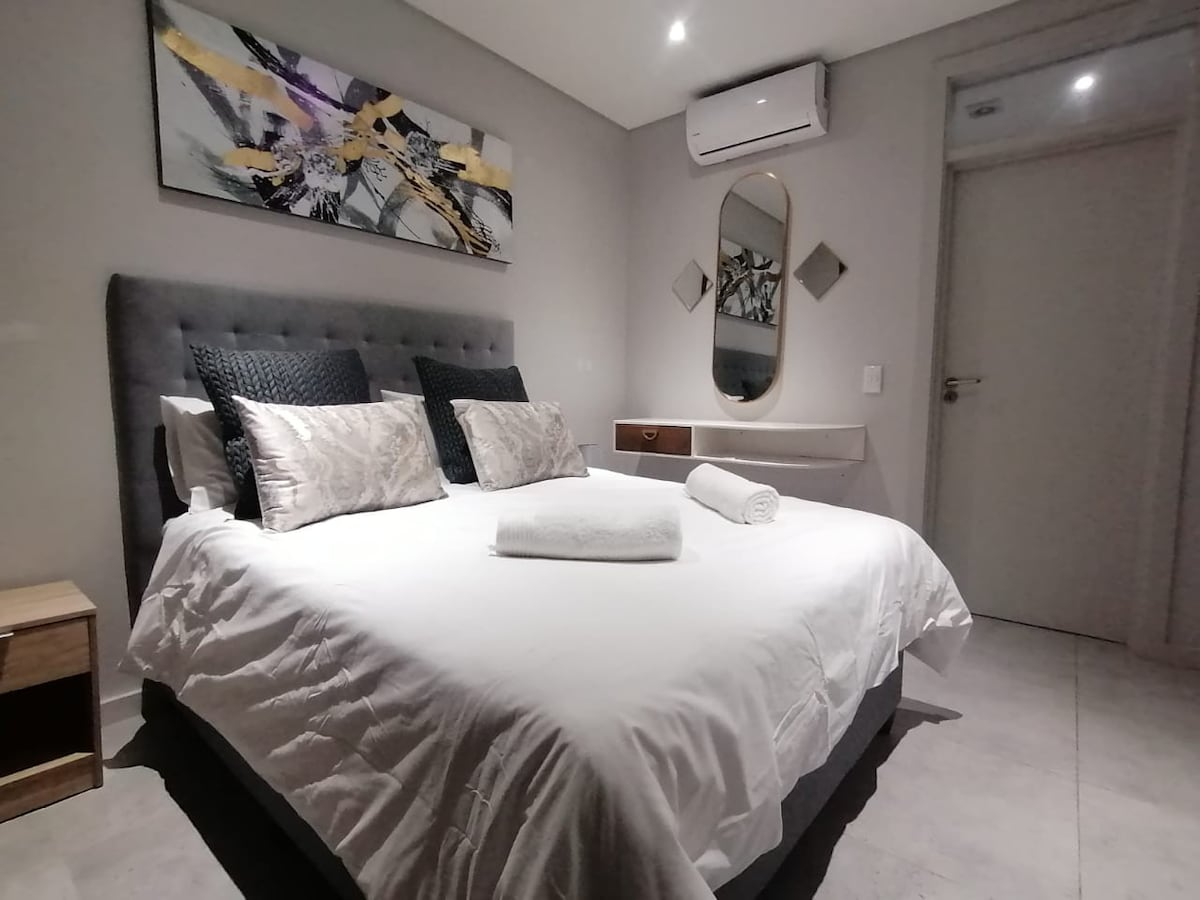 A modern bedroom features a spacious bed with neutral linens and decorative pillows. A piece of abstract wall art hangs above the bed. Natural light accentuates the space, and a bedside table sits to one side, providing functionality in a serene environment.