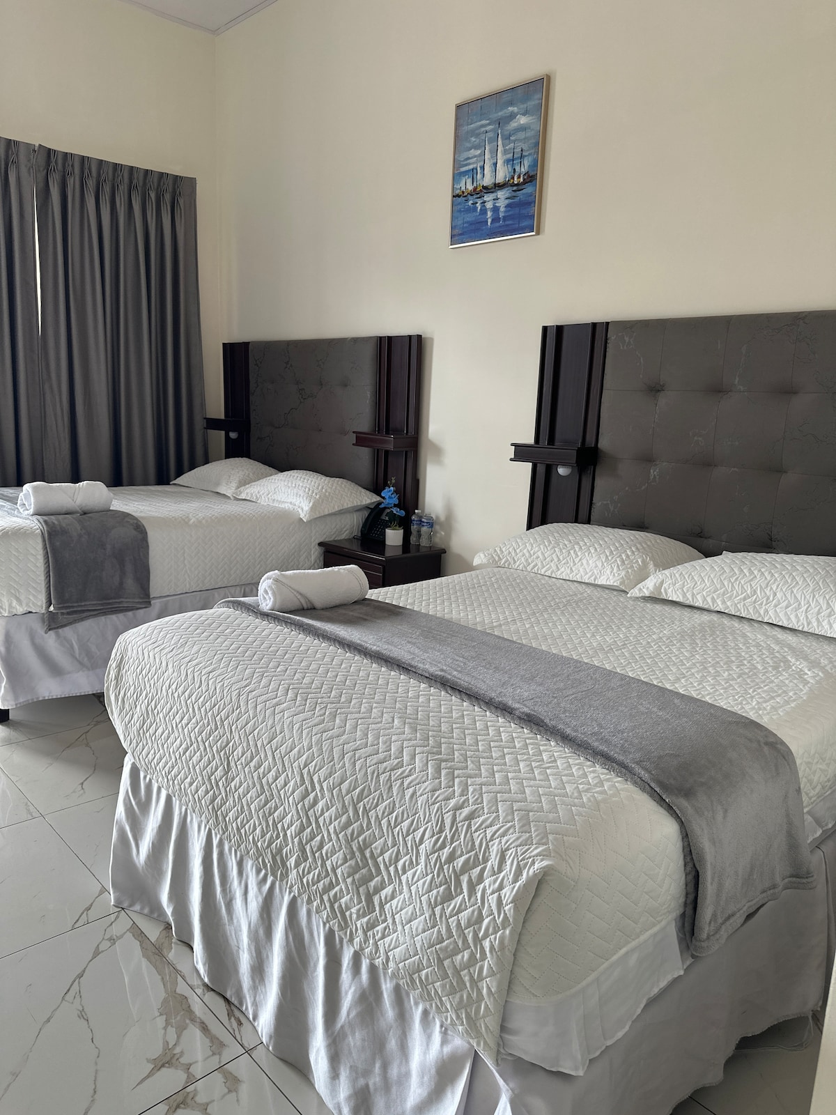 A spacious bedroom featuring two beds with upholstered headboards. Soft white linens cover the beds, complimented by light grey throws. A painting of a boat is mounted on the wall. Large grey curtains frame the window, and a small table with water bottles is placed beside the beds.