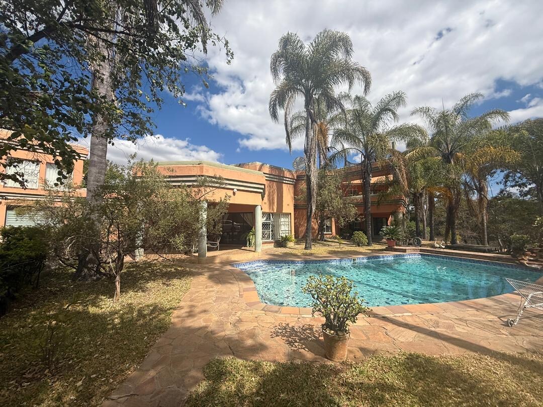 A serene outdoor space features a private pool surrounded by lush greenery and palm trees. The sunlit patio area shows a stone pathway leading to the pool, offering ample space for relaxation. The backdrop includes a spacious home with large windows, enhancing the inviting atmosphere.