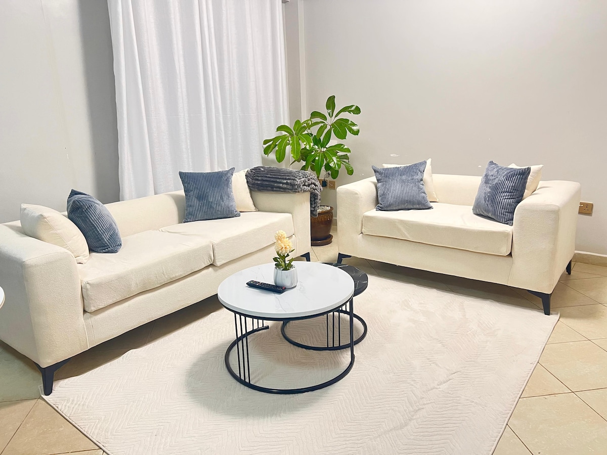 A bright living area features two cream-colored sofas adorned with blue accent pillows. A white coffee table, containing a small potted plant and remote control, occupies the center. Natural light filters through sheer curtains, enhancing the space's airy feel.