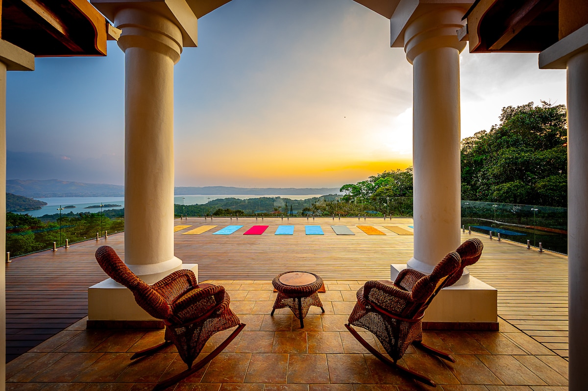 A serene view of a spacious deck is framed by two large columns, revealing a colorful yoga area and the lake beyond at sunset. Two wicker chairs and a small round table invite relaxation while the vibrant colors of the yoga mats harmonize with the natural landscape.