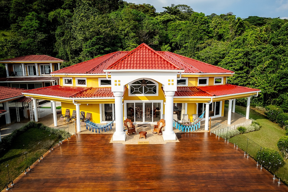 The villa features a vibrant yellow exterior complemented by a red tile roof. A spacious wooden deck surrounds the entrance, with several rocking chairs positioned for relaxation. Lush tropical greenery serves as a backdrop, enhancing the peaceful atmosphere of the property.