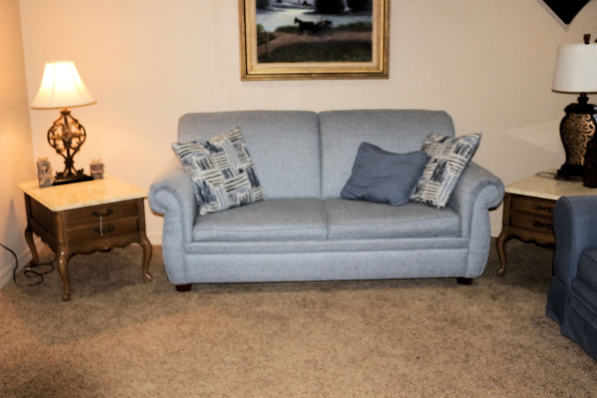 A light blue sofa is positioned in a cozy living area, complemented by two side tables with decorative lamps. Textured throw pillows enhance the sofa's comfort. A framed landscape painting adds a touch of decor to the warm ambiance of the room.