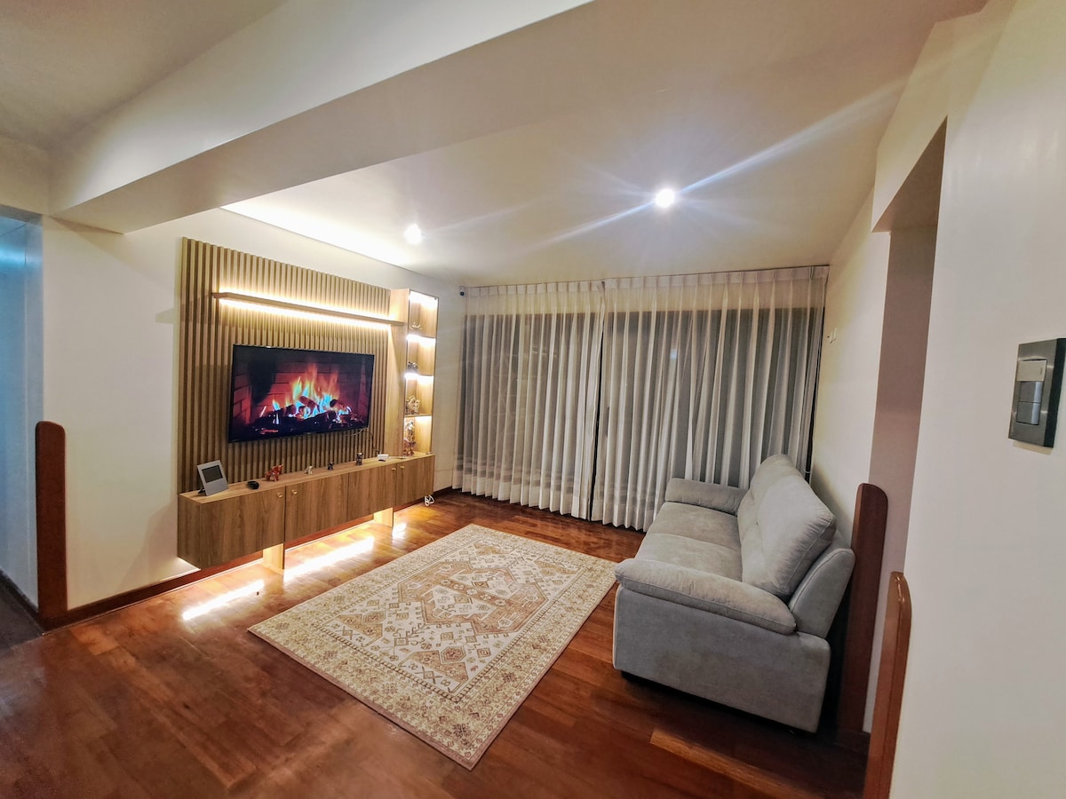 A spacious living area features a large, soft sofa positioned beside a decorative, wooden entertainment unit with an ambient mock fireplace. Natural light filters through vertical blinds covering wide windows, and a large area rug adds warmth to the hardwood flooring.