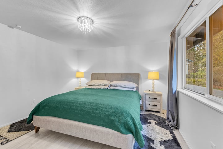 Una cama tamaño «king» con un cabecero tapizado en gris y un edredón con estampado floral en tonos verdes, amarillos y rosas. Cuatro almohadas descansan contra la cabecera. Las mesitas de noche a juego con las lámparas de mesa flanquean la cama, proyectando un brillo cálido y acogedor en la habitación.