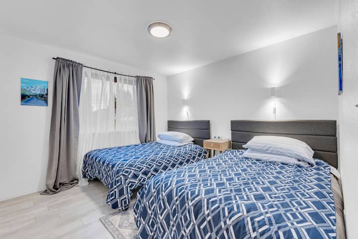 Un dormitorio luminoso con dos camas individuales bien hechas con ropa de cama geométrica azul. Las luces de pared suaves y una lámpara de techo crean un brillo acogedor. Una pequeña mesita de noche de madera se encuentra entre las camas y una gran ventana trae luz natural.
