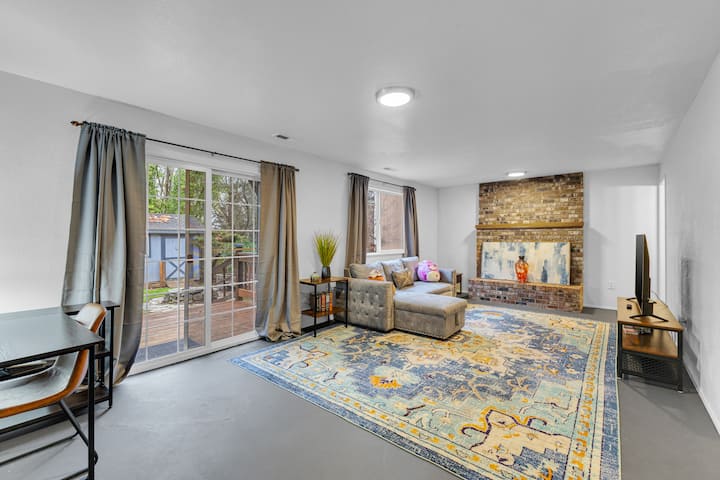 Una acogedora sala de estar con una gran alfombra azul estampada, un sofá cama seccional gris, una mesa auxiliar con libros y un soporte de TV. La chimenea de ladrillo cuenta con una pieza de arte moderno. Dos ventanas con cortinas dejan entrar la luz natural. La decoración incluye juguetes de peluche en el sofá.