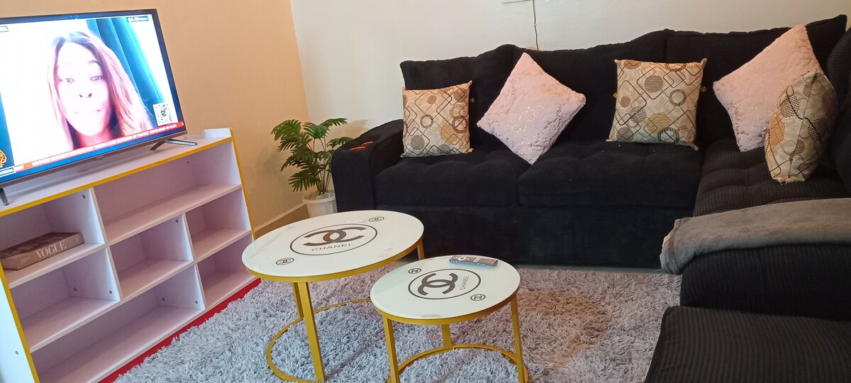 A modern living area features a dark sectional sofa adorned with decorative pillows. Two round tables with yellow accents are positioned on a soft rug. A television is mounted on a shelf, providing a clear central focus in the space.