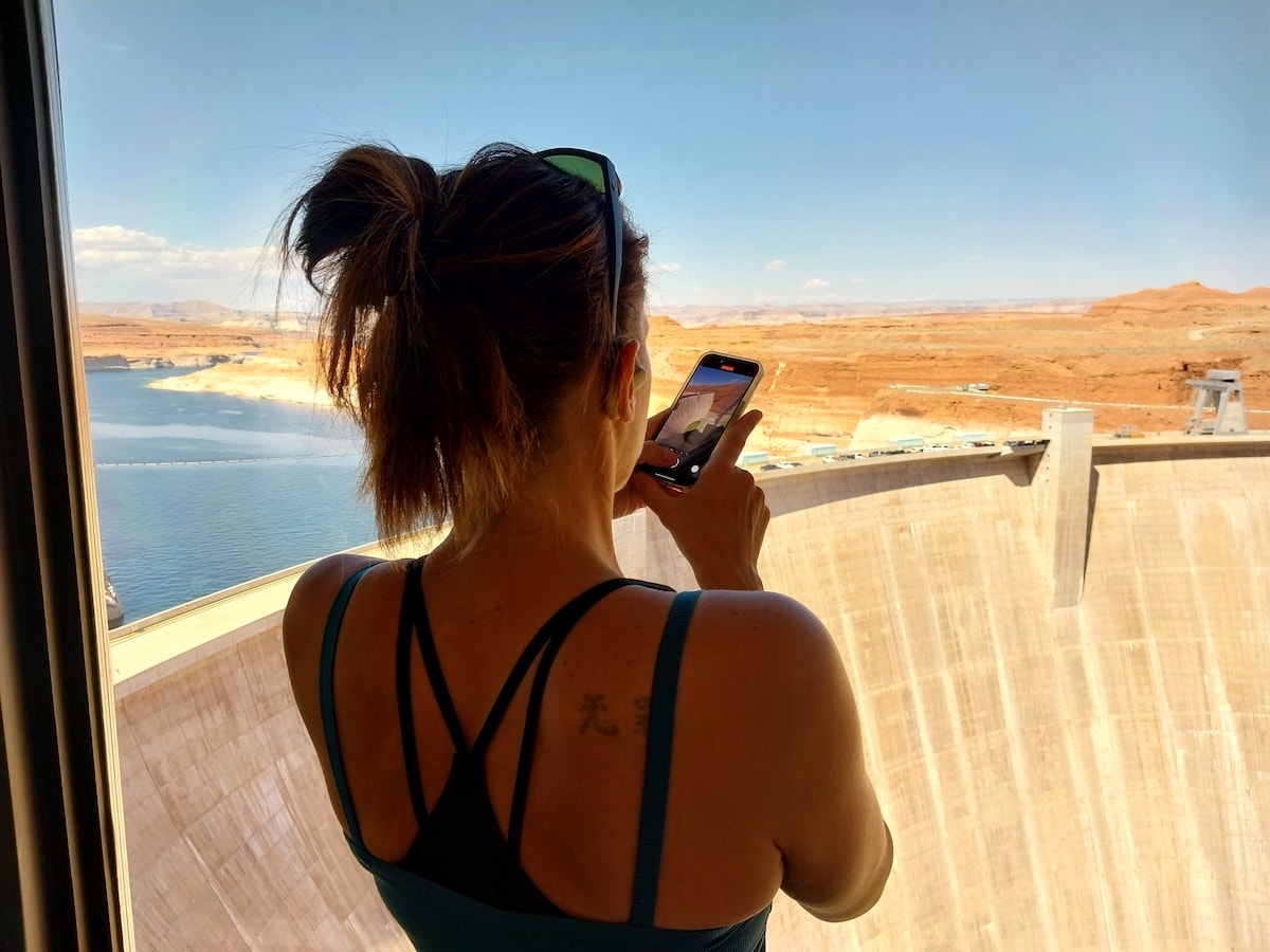 A person stands in silhouette at a large window, capturing the view of a vast landscape. The scene includes a body of water with rugged terrain in the background, under a clear blue sky with scattered clouds.