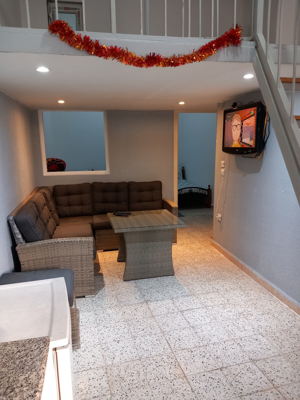 A cozy living area features a brown sectional couch arranged around a glass-topped table. Soft lighting is provided by recessed ceiling lights. A television is mounted on the wall, and a charming decorative garland is secured along the upper edge, adding a festive touch.