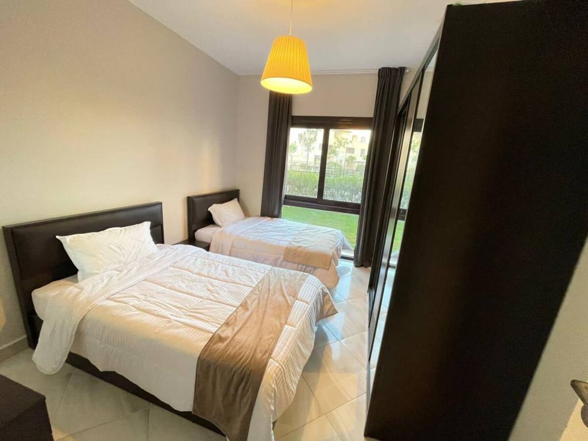 A well-lit bedroom features two beds, one spacious and one smaller, both dressed in white linens. A large window offers a view of the greenery outside, while dark curtains frame the window. A minimalist wardrobe stands against one wall, adding to the serene ambiance.