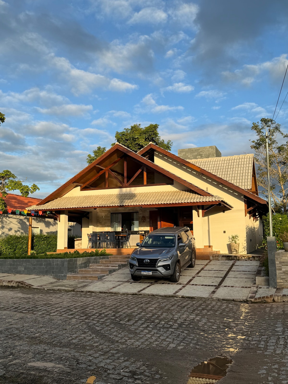 The exterior of the house features a triangular roof with wooden accents, set against a backdrop of blue skies and scattered clouds. A vehicle is parked in front on a stone pathway, flanked by manicured greenery and a welcoming entrance.