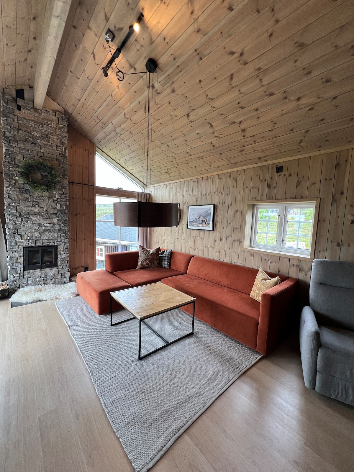 A spacious living area features a corner sofa in warm tones, paired with a simple coffee table. Natural light floods the room through large windows, casting a soft glow on wooden walls and floors, while a decorative fireplace adds to the inviting ambiance.