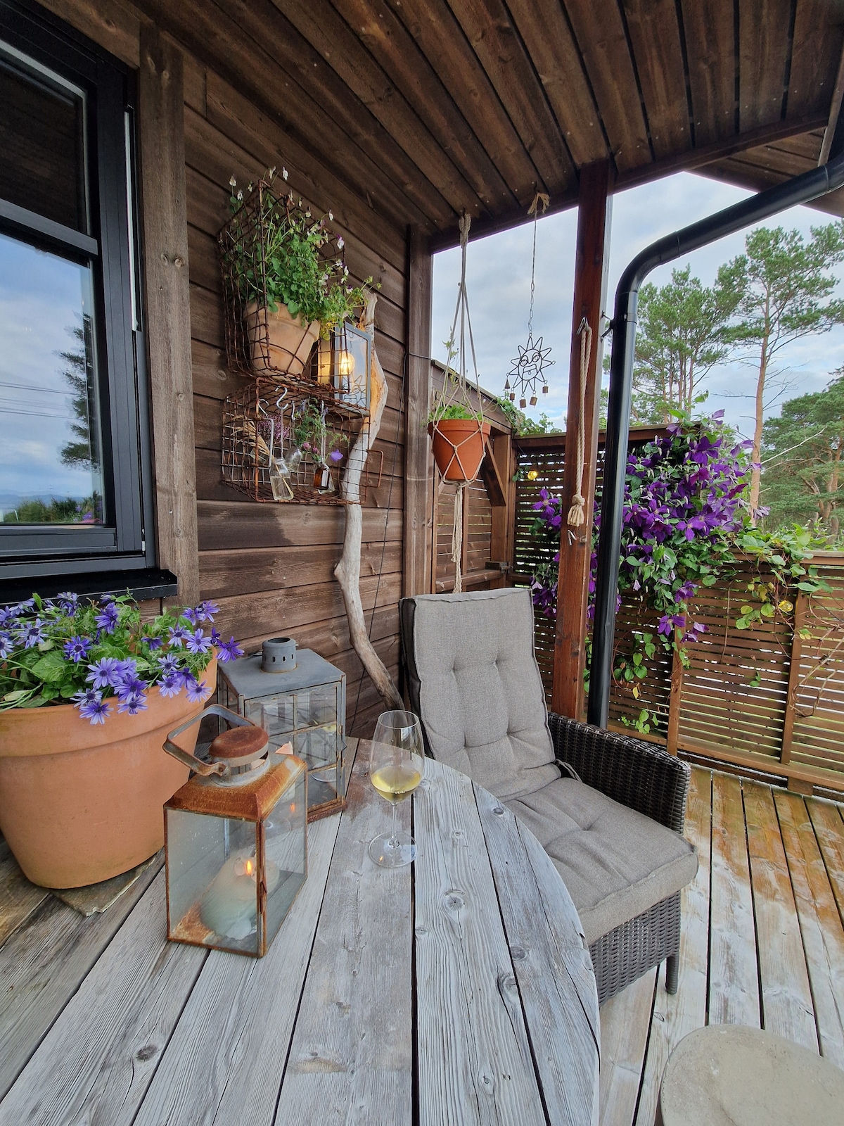 A cozy terrace area featuring a wooden table and a comfortable chair, surrounded by potted plants and vibrant purple flowers. A lantern and a glass of wine add a tranquil touch, while wooden panels create a serene outdoor space.