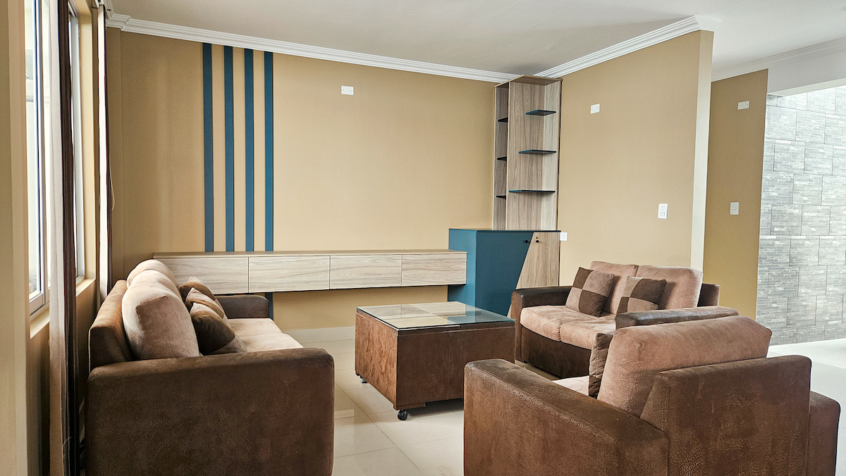 A spacious living area is presented, featuring multiple comfortable brown sofas arranged around a glass coffee table. A light brown accent wall with blue stripes adds character, while cabinetry provides functional storage. Natural light fills the room through large windows.
