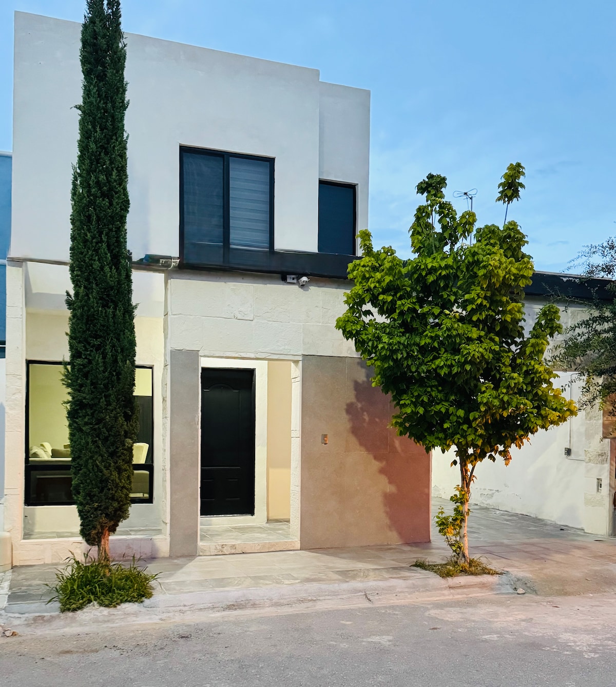 The exterior of the house features a modern design with a smooth façade and a prominent black front door. Tall trees frame the entrance, surrounded by a clean, paved pathway leading to the home. Large windows allow natural light to enhance the inviting atmosphere.