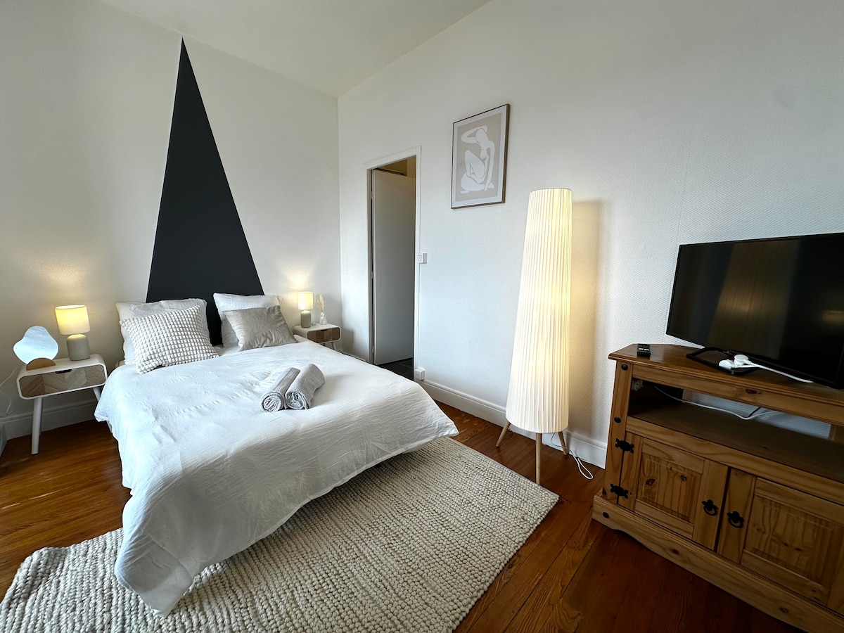 The cozy sleeping area features a spacious bed with soft linens, complemented by decorative cushions. A large window invites natural light, while two bedside lamps offer gentle illumination. A TV is positioned on a wooden cabinet, and a modern floor lamp adds warmth to the ambiance.