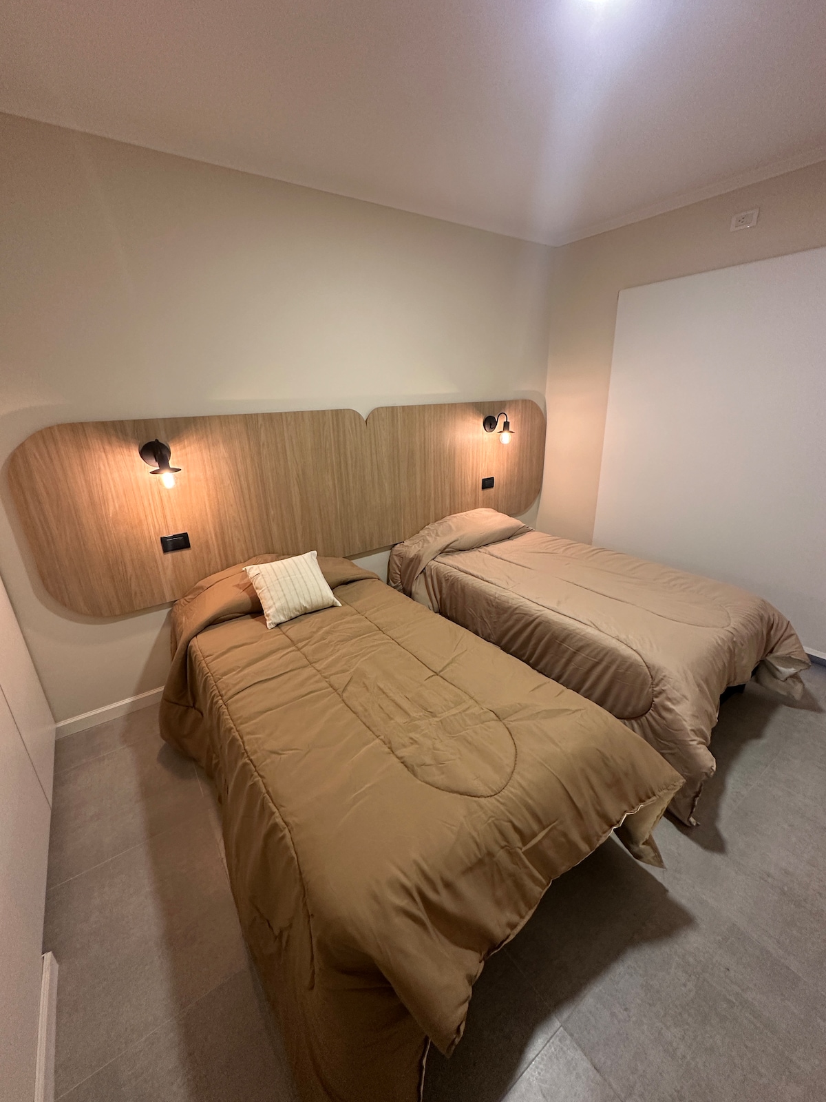 A well-lit bedroom features two neatly made beds with beige bedding against a wooden headboard. Simple wall sconces add soft illumination, while a textured cushion is placed on one bed. Light-colored walls and a minimalist design create a serene ambiance.