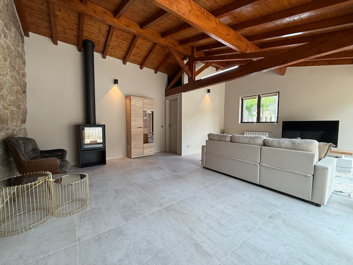 A spacious living area features exposed wooden beams and a stone wall, complemented by a modern fireplace and large windows allowing natural light. A light-colored sofa and a pair of chairs provide ample seating, alongside a decorative side table, creating a comfortable environment.