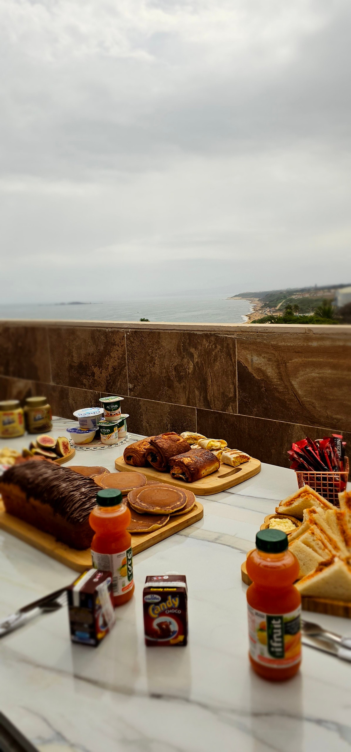 A breakfast spread is arranged on a table featuring pastries, breads, and drinks. The ocean view in the background adds a scenic element to the setting. Various food items are presented alongside containers and utensils, creating an inviting atmosphere for a relaxed morning.